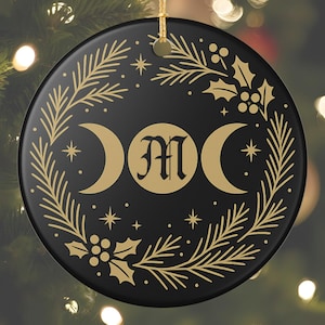 May include: Black ceramic Christmas ornament with a gold triple moon design, the letter "M", stars, and a botanical wreath. The ornament hangs from a gold string.
