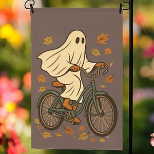 May include: A garden flag depicting a cartoon ghost on a teal bicycle. The ghost is draped in a white sheet, with orange and yellow autumn leaves scattered around. The background is a muted brown.