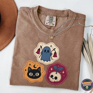 Könnte beinhalten: Braunes T-Shirt mit drei gestickten Aufnähern: ein blauer Geist, eine schwarze Katze mit Mond und ein Totenkopf mit einer Pflanze. Eine silberne Halskette und ein brauner Hut sind ebenfalls sichtbar. Das Shirt hat ein Comfort Colors-Etikett.