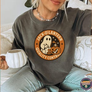 May include: Dark gray t-shirt with a circular graphic featuring a ghost, pumpkin, and the text "Jack-O-Lantern Quality Control." The shirt is part of a Halloween-themed collection. A white pumpkin-shaped mug is held in the image.