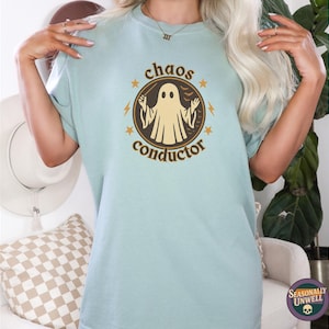 May include: A light blue t-shirt with a circular graphic featuring a ghost figure and the words "chaos conductor". The design is in brown and gold tones. The shirt is a comfortable, casual style.