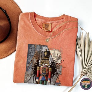 May include: Coral-colored t-shirt featuring a graphic of a nutcracker soldier. The shirt has a crew neck and a tag that reads "Comfort Colors". A gold necklace and a brown hat are also in the image. The shirt is part of a clothing collection.