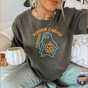 May include: Dark gray t-shirt with a neon graphic of a ghost holding a knife and a pumpkin, with the text "PUMPKIN CARVER". The shirt is part of a Halloween-themed collection.
