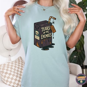 May include: Light blue t-shirt featuring a graphic of a carton labeled "Tears of My Enemies." The carton is dark brown with a skull and dripping liquid design. A straw and a tag that says "DEAD CONTENTS" are also visible.