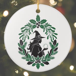 Könnte beinhalten: Runder weißer Keramik-Weihnachtsschmuck mit einer schwarzen Hexensilhouette, die eine Kristallkugel hält, umgeben von einem grünen Stechpalmen- und Mistelzweigkranz. Die Hexe trägt einen spitzen Hut. Ein goldener Faden ist zum Aufhängen befestigt.