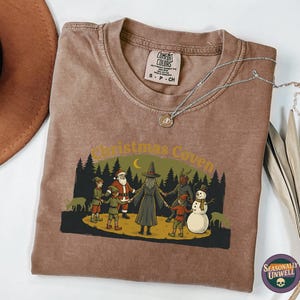 May include: Brown t-shirt with a graphic featuring a witch, Santa Claus, and other characters in a forest setting. The text "Christmas Coven" is arched above the scene. A silver necklace with a pendant is on the shirt.