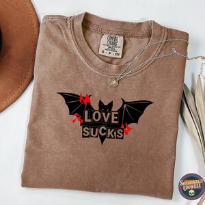 May include: Brown t-shirt with a black bat graphic and the words "LOVE SUCKS". The shirt has a crew neck and short sleeves. A silver necklace with a pendant is on the shirt. A brown hat is visible in the background.