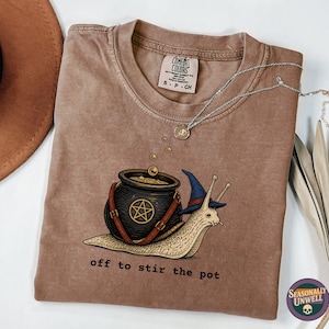 Może przedstawiać: Brązowy t-shirt z krótkim rękawem z grafiką ślimaka w niebieskim kapeluszu czarownicy, niosącego czarny kociołek z symbolem pentagramu. Poniżej grafiki znajduje się napis "off to stir the pot". Koszulka posiada metkę marki Comfort Colors.