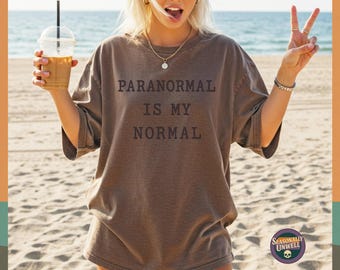 May include: Brown t-shirt with the text "PARANORMAL IS MY NORMAL" in a dark brown font. The shirt is worn on a beach with a drink in hand. The shirt is from Seasonally Unwell.