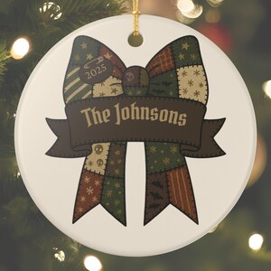May include: A round, white Christmas ornament with a patchwork bow design. The bow features various patterns in green, brown, and red. A banner across the bow reads "The Johnsons" and the year "2025" is also displayed.