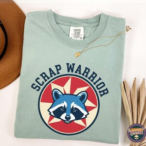 Peut inclure: T-shirt vert clair avec l'inscription "SCRAP WARRIOR" entourant un dessin de raton laveur. Le raton laveur est bleu et blanc, sur un motif d'étoile rouge et blanche. Un chapeau marron et un collier doré sont visibles.