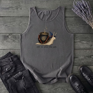 May include: A charcoal gray sleeveless tank top featuring a graphic of a snail wearing a blue wizard hat and carrying a black cauldron with a pentagram symbol. The text below the graphic reads, off to stir the pot, in a simple black font.