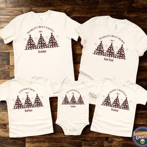 以下が含まれることがあります： クリスマスをテーマにしたグラフィックがプリントされた、オフホワイトのTシャツとロンパースのコレクションです。「World's Best Santa」や「Merry Crispmas」などのテキストと家族の名前がプリントされています。グラフィックにはクリスマスツリーが含まれています。
