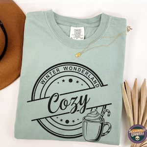 May include: A light green Comfort Colors t-shirt with a black graphic that says "Winter Wonderland Cozy" with a mug of hot chocolate. A brown hat and a gold necklace are visible in the background.