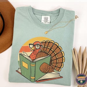 May include: A light blue Comfort Colors t-shirt featuring a cartoon turkey wearing glasses and reading a book. The turkey is set against a retro sunset design. The shirt has the text "Seasonally Unwell" on the bottom right.