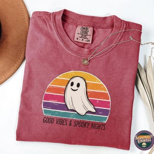 Könnte beinhalten: Rotes T-Shirt mit einer Grafik eines lächelnden Geistes vor einem Sonnenuntergangsdesign. Der Text "GOOD VIBES & SPOOKY NIGHTS" befindet sich darunter. Eine silberne Halskette mit Anhänger liegt auf dem Shirt.