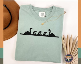 May include: A light green t-shirt with a black graphic of the Loch Ness Monster and waves. The shirt has a crew neck and short sleeves. A gold necklace and a brown hat are also in the image.