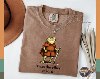 May include: Brown t-shirt featuring a cartoon frog in a cape, holding a sword, with the text "'twas the vibes m'lord". A silver necklace and a brown hat are also visible. The t-shirt is a Comfort Colors brand.