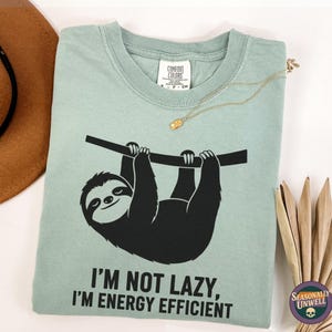May include: A pale green t-shirt featuring a black sloth design hanging from a branch. The text reads "I'M NOT LAZY, I'M ENERGY EFFICIENT". A brown hat and gold necklace are visible in the background.
