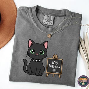May include: A gray t-shirt featuring a cartoon black cat with green eyes and a studded collar. The cat sits next to a small chalkboard that reads "100 lessons". A silver necklace and a brown hat are also visible.