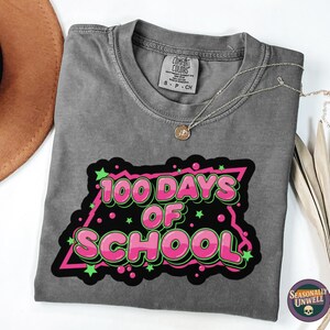 May include: Grey t-shirt featuring the text "100 Days of School" in pink and green. The shirt has a crew neck and short sleeves. A gold necklace and a brown hat are also in the image.