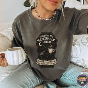 May include: Dark gray t-shirt with a coffin-shaped graphic that reads "Coven of the Perpetually Tired." The graphic includes a crescent moon, coffee cup, and a sleeping cat. The text "Fueled by caffeine, held together by sarcasm" is also present.