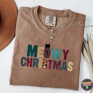 May include: A brown t-shirt with the words "MEOWY CHRISTMAS" in a patchwork design. The letters are in various colours and patterns, including plaid and floral. A silver necklace and a brown hat are also visible.