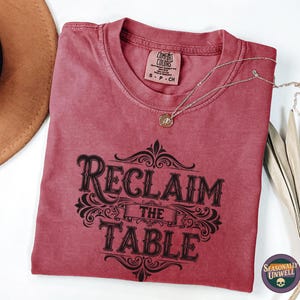 May include: A red t-shirt with the text "Reclaim the Table" in a decorative font. A gold necklace and a brown hat are also visible. The shirt has a small tag that says "Comfort Colors".