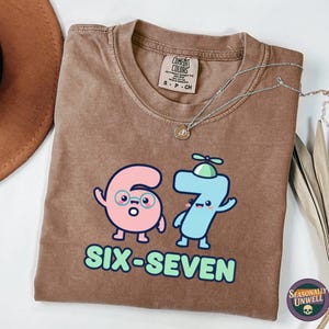 Può includere: T-shirt marrone con una grafica dei numeri sei e sette come personaggi dei cartoni animati, con le parole "SIX-SEVEN" sotto. La maglietta ha un girocollo e maniche corte. Una collana d'argento con un piccolo ciondolo è sulla maglietta.