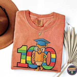 May include: Coral-coloured t-shirt with a colourful graphic of a pencil wearing a graduation cap and glasses, in front of the number 100. A brown hat and gold necklace are also visible.