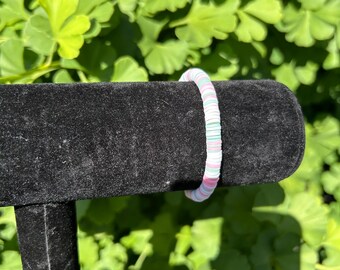 May include: A colorful bracelet with a flat, disc-like bead design. The bracelet features a mix of white, pink, and light green beads. The bracelet is displayed on a black velvet jewelry stand against a backdrop of green foliage.