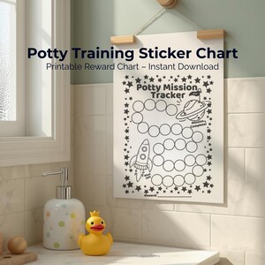Puede incluir: Un gráfico de pegatinas para el entrenamiento para ir al baño con el texto "Potty Mission Tracker" e ilustraciones de un cohete y un planeta. El gráfico está colgado en una pared cerca de una ventana, un dispensador de jabón y un pato de goma amarillo.