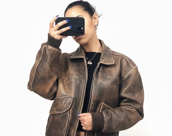 May include: Brown leather bomber jacket with a distressed finish. The jacket features a zip-up front, two front pockets, and ribbed cuffs and hem. The jacket is worn with a black top and light-colored skirt.
