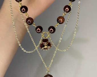 Peut inclure: Un collier multi-rangs composé d'une rangée de perles sphériques marron foncé reliées par des chaînes en métal doré. Le bijou présente une petite breloque en forme de tasse à thé dorée et un pendentif ovale lisse de couleur marron foncé.