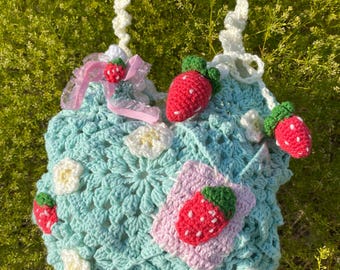 May include: A handmade crochet bag in light blue, adorned with red strawberry appliqués and white flower accents. The bag features a white strap and a pink ribbon with a small strawberry. The bag is a unique, whimsical accessory.