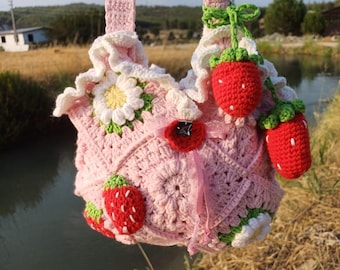 May include: A handmade pink crochet bag with a strawberry theme. The bag features crocheted strawberries, white daisies, and a red button closure. The bag has two pink straps and is set against a natural outdoor background.