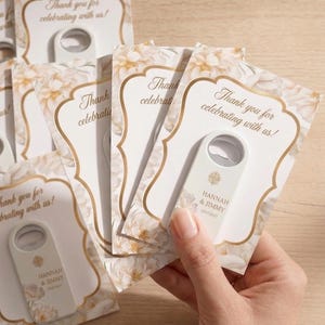 May include: A collection of bottle openers with floral design cards. Each card has a gold border and the text "Thank you for celebrating with us!". The bottle openers are white with floral accents and the names "HANNAH & JIMMY".