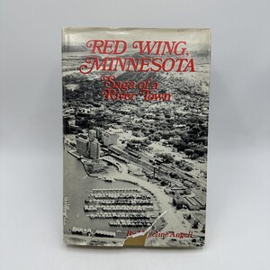 Puede incluir: Un libro antiguo titulado "Red Wing, Minnesota, Saga of a River Town" con una fotografía aérea en blanco y negro de una ciudad y un río. El título está en escritura roja. El libro tiene una cubierta desgastada.