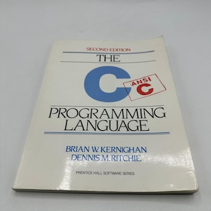 Könnte beinhalten: Ein weißes Buch mit dem Titel "The C Programming Language" von Brian W. Kernighan und Dennis M. Ritchie. Das Cover zeigt ein großes blaues "C" und schwarzen Text. Das Buch ist eine zweite Auflage und hat einen roten "ANSI C" Stempel.
