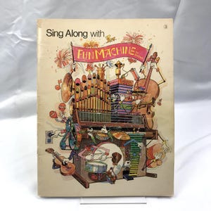 May include: A colorful book cover with the title "Sing Along with Fun Machine" by  "The  Bee Gees". The cover features a cartoon illustration of a musical instrument with various musical instruments and characters playing them.