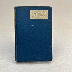 Könnte beinhalten: Ein Vintage-Buch mit dunkelblauem Einband. Der Titel "THE PLAYS OF J. M. BARRIE" ist auf einem rechteckigen Etikett auf der Vorderseite aufgedruckt. Das Buch ist leicht abgenutzt, was auf Alter und Nutzung hindeutet. Der Hintergrund ist eine schlichte weiße Oberfläche.