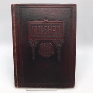 Könnte beinhalten: Ein dunkelweinrotes Hardcover-Buch mit strukturierter Oberfläche. Der Vorderdeckel zeigt einen geprägten rechteckigen Rahmen mit dem Text KULSHAN in Großbuchstaben über einem kreisförmigen Emblem, in dessen Mitte das Jahr 1928 eingeprägt ist.