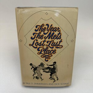May include: A vintage book with the title "The Year The Mets Lost Last Place." The cover displays a baseball diamond design with the title in blue and orange script. Illustrations of baseball players are below the title. The authors' names are at the bottom.