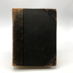 May include: An antique book with a worn, black leather cover. The edges of the cover are brown and show signs of wear. The spine is also black, with a vertical line down the center. The book's corners are slightly rounded.