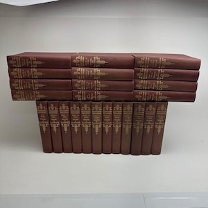 May include: A collection of maroon Encyclopedia Britannica books arranged in four horizontal rows and one vertical row. Each spine features gold lettering and decorative wheat-like symbols. The text includes volume numbers and alphabetical ranges.