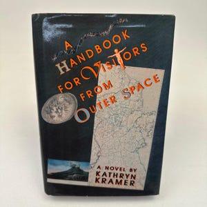 May include: A Handbook for Visitors from Outer Space book cover. The cover is dark blue with orange lettering and illustrations. The book features a map, a seashell, and a small illustration of a person. The author's name, Kathryn Kramer, is at the bottom.