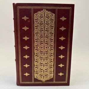 May include: A maroon hardcover book featuring a central vertical panel with intricate gold filigree patterns. The cover is framed by thin gold lines and decorated with small, repeating gold floral motifs on both the left and right sides.
