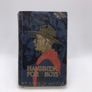 Op de afbeelding: Vintage boek "Handbook for Boys" met een blauwe kaft met een profiel van een man met een hoed en rode sjaal. De titel van het boek staat in gouden letters, met daaronder "Boy Scouts of America".