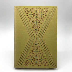 Op de afbeelding: Een hardcover boek met een lichtgele kaft. De kaft heeft een symmetrisch ontwerp met rode, groene en blauwe bloempatronen en diagonale lijnen. Het ontwerp van het boek doet denken aan vintage of antieke stijlen.