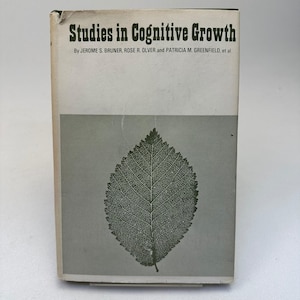 May include: A hardcover book titled Studies in Cognitive Growth by Jerome S. Bruner, Rose R. Olver, and Patricia M. Greenfield, et al. The cover features a white top section with black text and a gray bottom section with a detailed black leaf illustration.